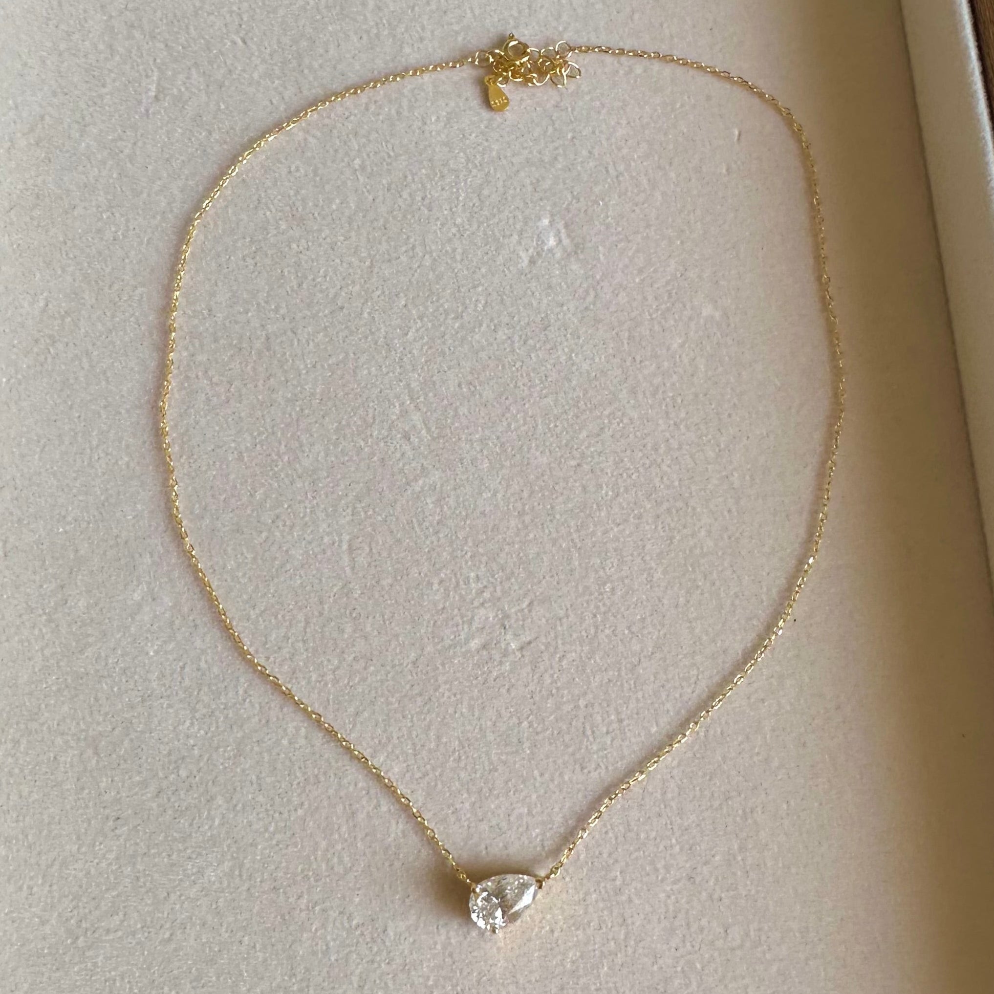A delicate gold chain necklace with a small clear teardrop-shaped gemstone pendant, displayed inside a cream-colored jewelry box.