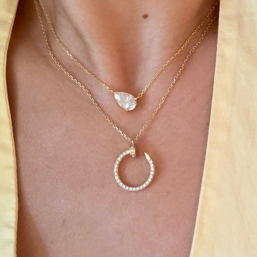 A close-up of a person's neck and chest wearing a layered gold necklace set. The top layer features a delicate chain with a small, irregular clear gemstone pendant, while the bottom layer has a larger gold crescent moon pendant encrusted with small clear stones.