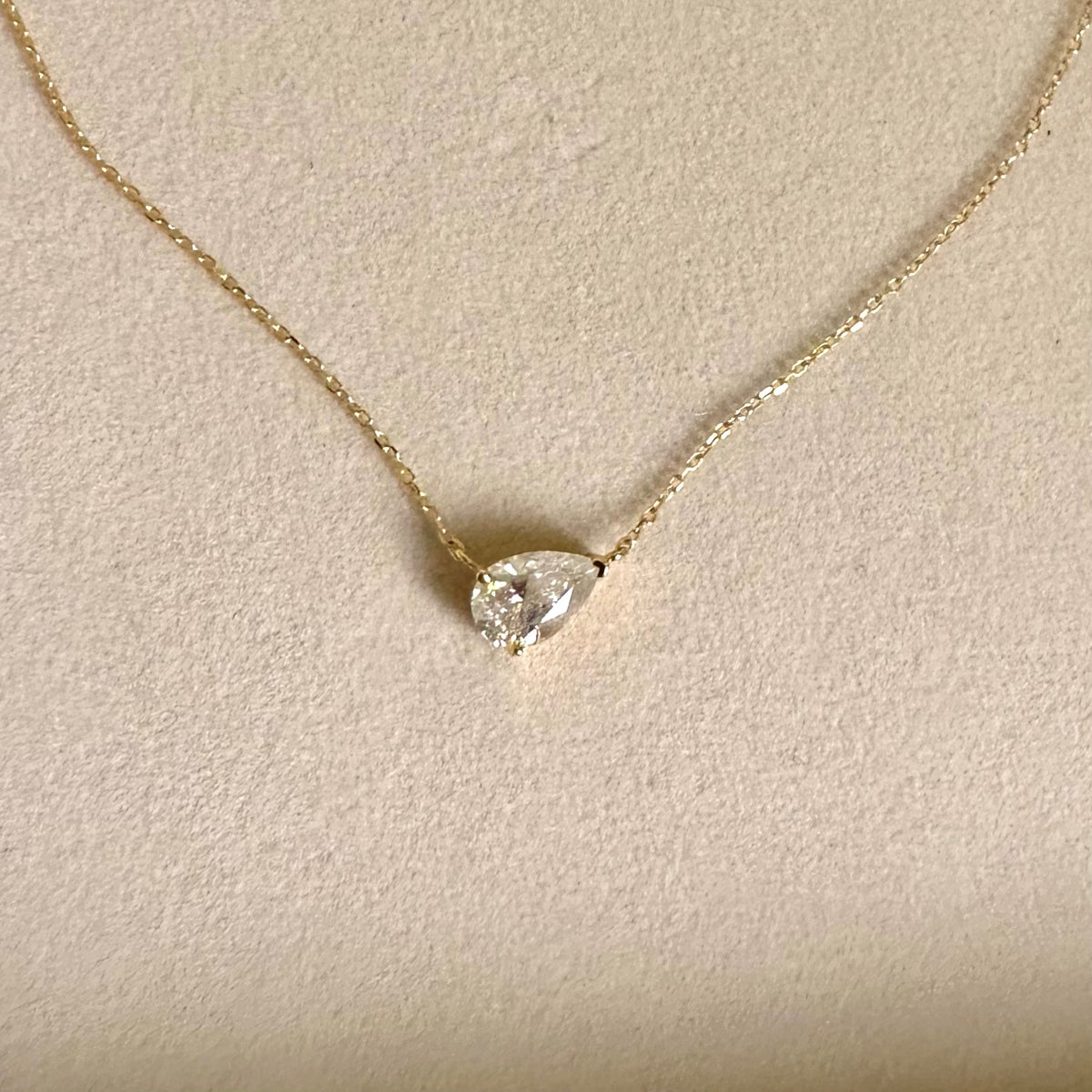 A delicate gold chain necklace with a pear-cut clear gemstone pendant, displayed on a textured beige surface.